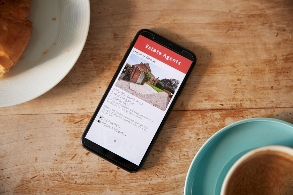 Close Up Of Mobile Phone With Realtors Property App Lying On Café Table